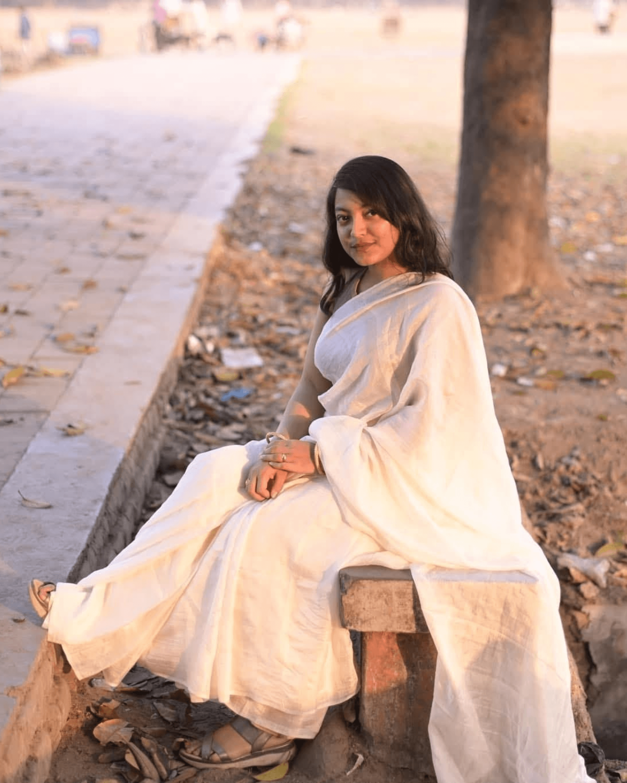 Young woman wearing a white saree, sitting outdoors on a stone bench with a serene expression.